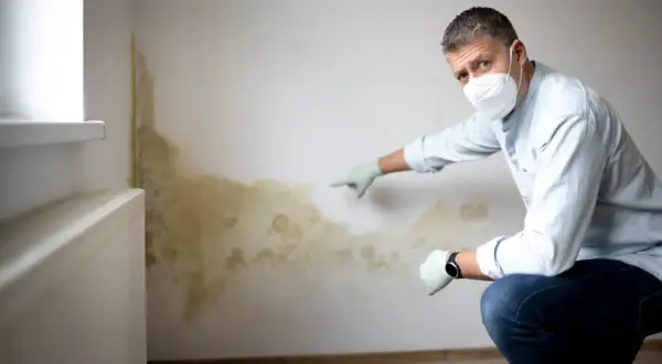 Mann mit Maske und Handschuhen zeigt auf deutlichen **Schimmel an der Wand**, der sich neben einem Heizkörper ausgebreitet hat.