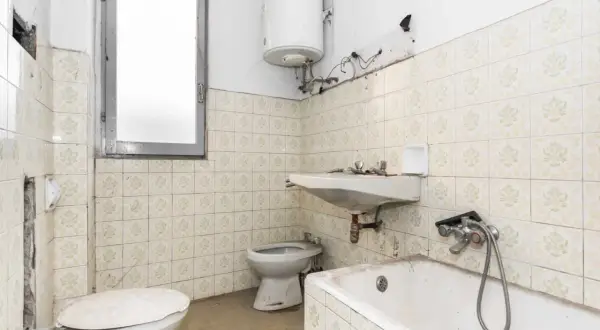 Badezimmer mit veralteten Fliesen, bereit zum Streichen. Toilette, Waschbecken und Badewanne sind sichtbar.