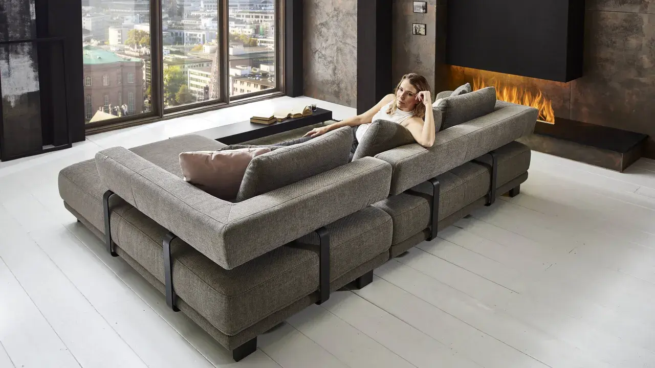 Moderne Wohnlandschaft mit grauem Stoffbezug, die als **Sofa als Raumteiler Ideen** dient. Eine Frau entspannt sich darauf, im Hintergrund ein Kaminfeuer.