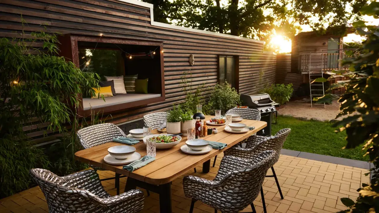 Eine schmale Terrasse gestalten: Gemütlicher Essbereich mit Holztisch, Rattanstühlen, Grill und Sitznische mit Kissen.
