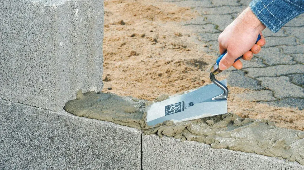 Maurer trägt Mörtel mit Kelle auf Betonsteine auf. Der Unterschied zwischen Beton und Zement wird hier beim Bau deutlich.