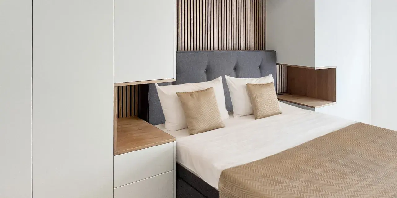 Ein gemütliches kleines Schlafzimmer einrichten mit einem Bett, grauem Kopfteil, weißen und goldenen Kissen, Holzakzenten und weißen Schränken.
