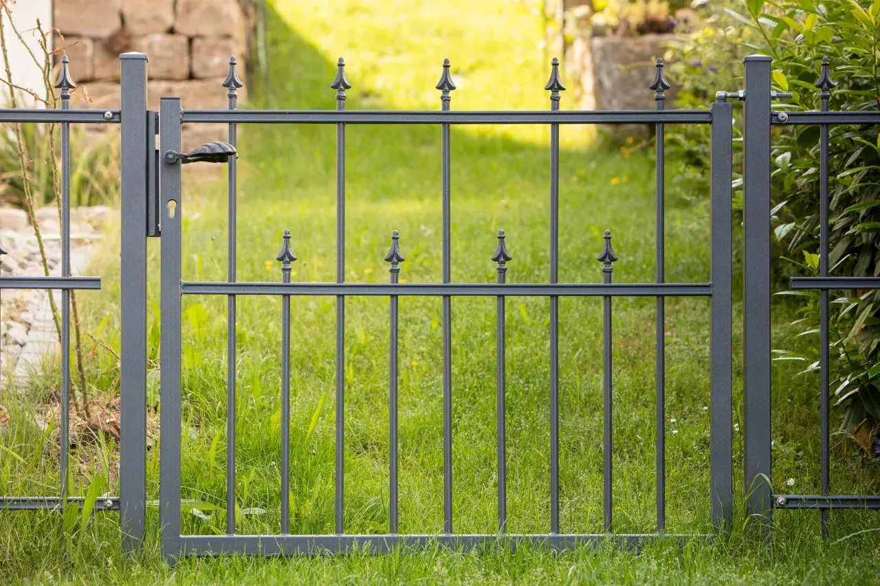 Graues Metallgartentor mit Griff und Schloss, bereit zum **Gartentor einbetonieren**. Dahinter grüner Rasen und eine Steinmauer.