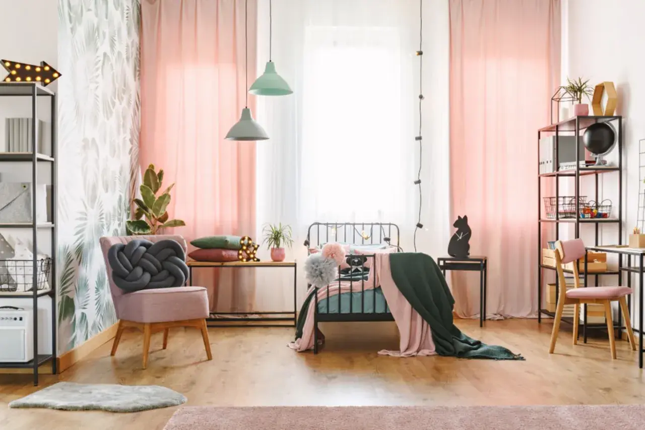Ein stilvolles Jugendzimmer einrichten: Rosa Vorhänge, ein Bett mit Decken, ein Sessel mit Knoten-Kissen und ein Schreibtisch mit Regal.