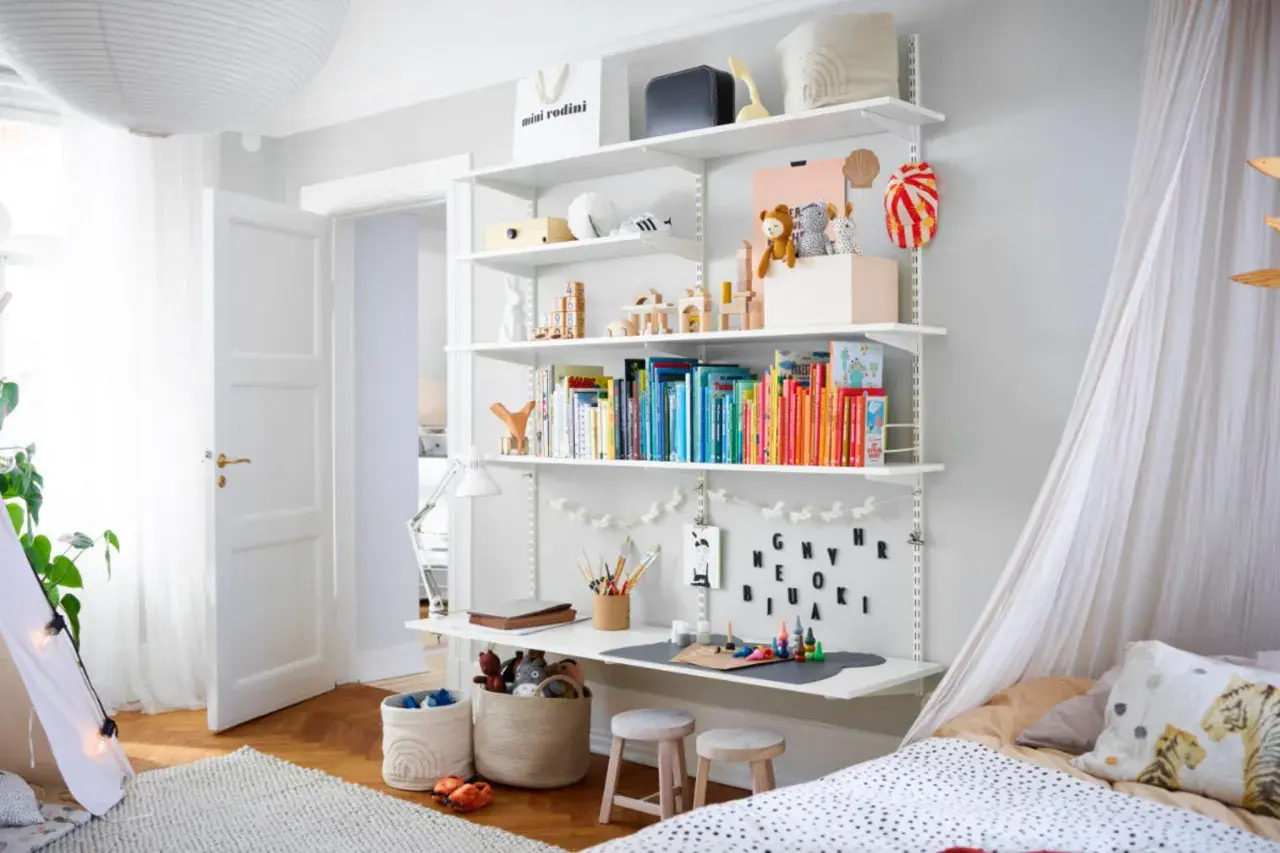 Ein schmal eingerichtetes Kinderzimmer mit einem Regal voller Bücher und Spielzeug, einem Schreibtisch und einem Bett mit Baldachin.