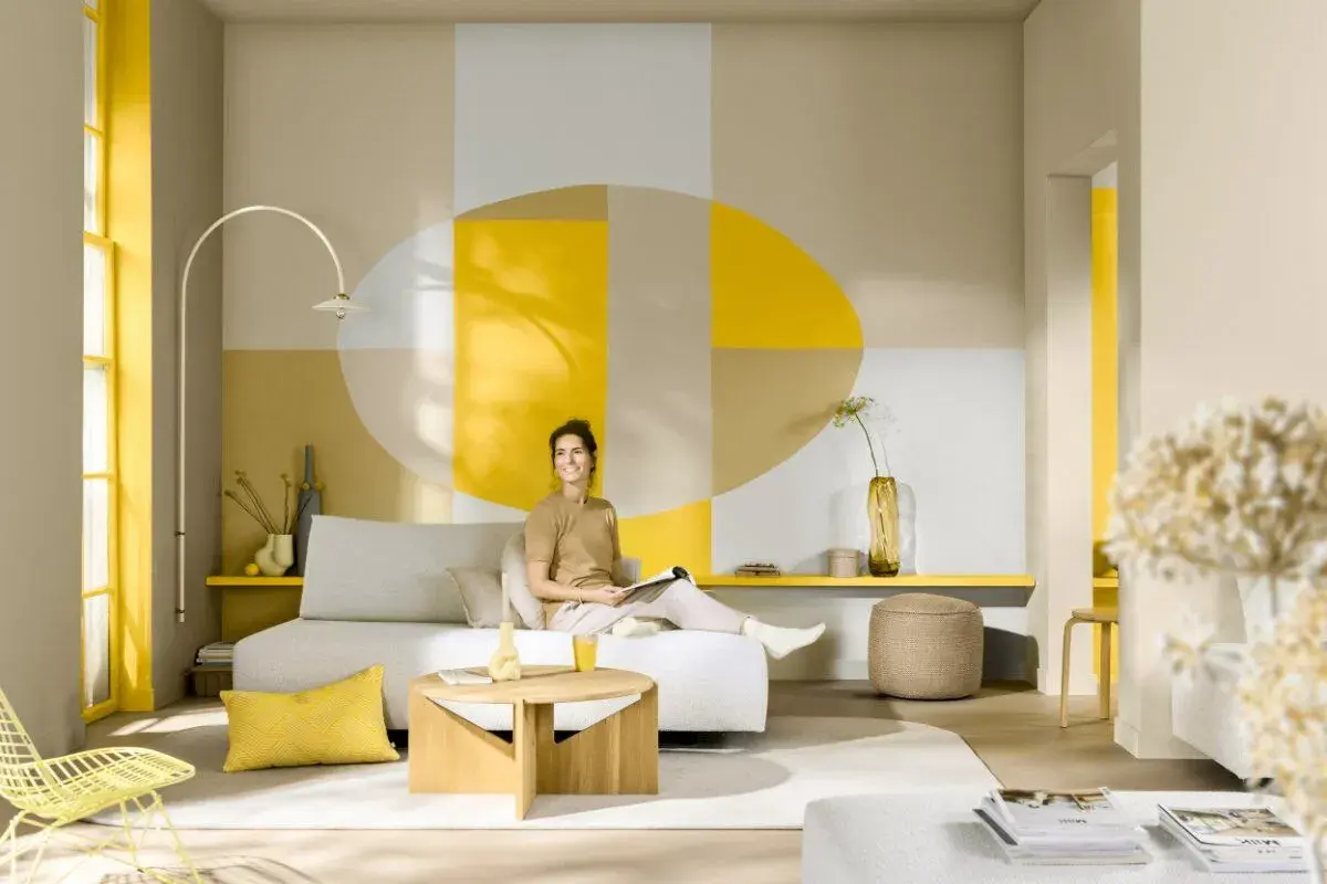 Eine Frau entspannt sich in einem hellen Wohnzimmer mit geometrischen Wandmustern. Die Frage, welche Farbe passt zu Gelb, wird hier mit Beige und Grau beantwortet.