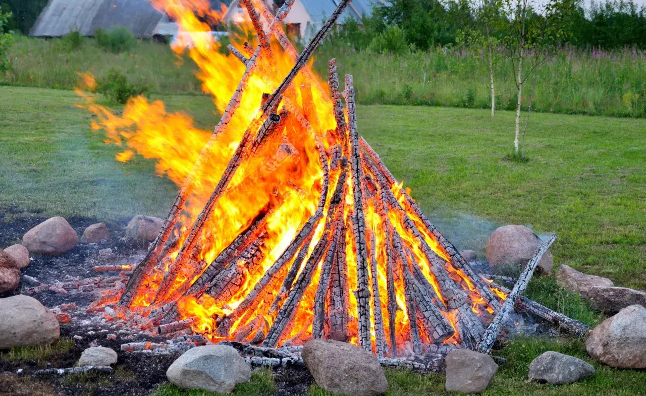 Ein großes, loderndes lagerfeuer im garten, umgeben von Steinen. Die Flammen tanzen hoch und erhellen die grüne Wiese.