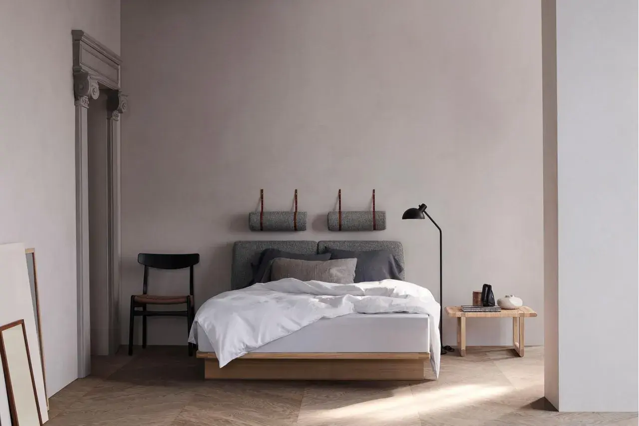 Ein helles, skandinavisches Schlafzimmer mit einem Holzbett, grauen Kissen und einer Stehlampe. Zwei gerollte Decken hängen an der Wand.