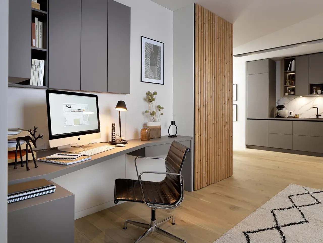 Moderne homeoffice ideen: Ein aufgeräumter Schreibtisch mit iMac, Lederstuhl und Regalwand. Daneben eine Holzwand, die in eine Küche übergeht.