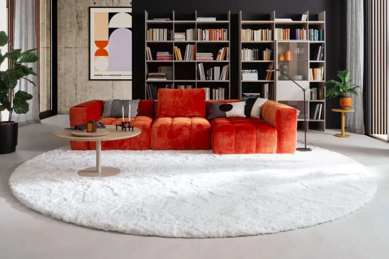 Ein flauschiger, weißer Teppich liegt unter dem auffälligen, orangefarbenen Sofa und dem runden Couchtisch.