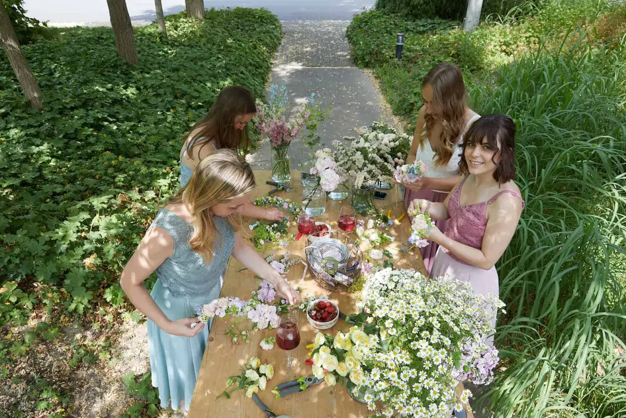 Vier Frauen binden im Freien einen Blumenkranz. Sie sitzen an einem Holztisch voller Blumen, Werkzeuge und Getränke.