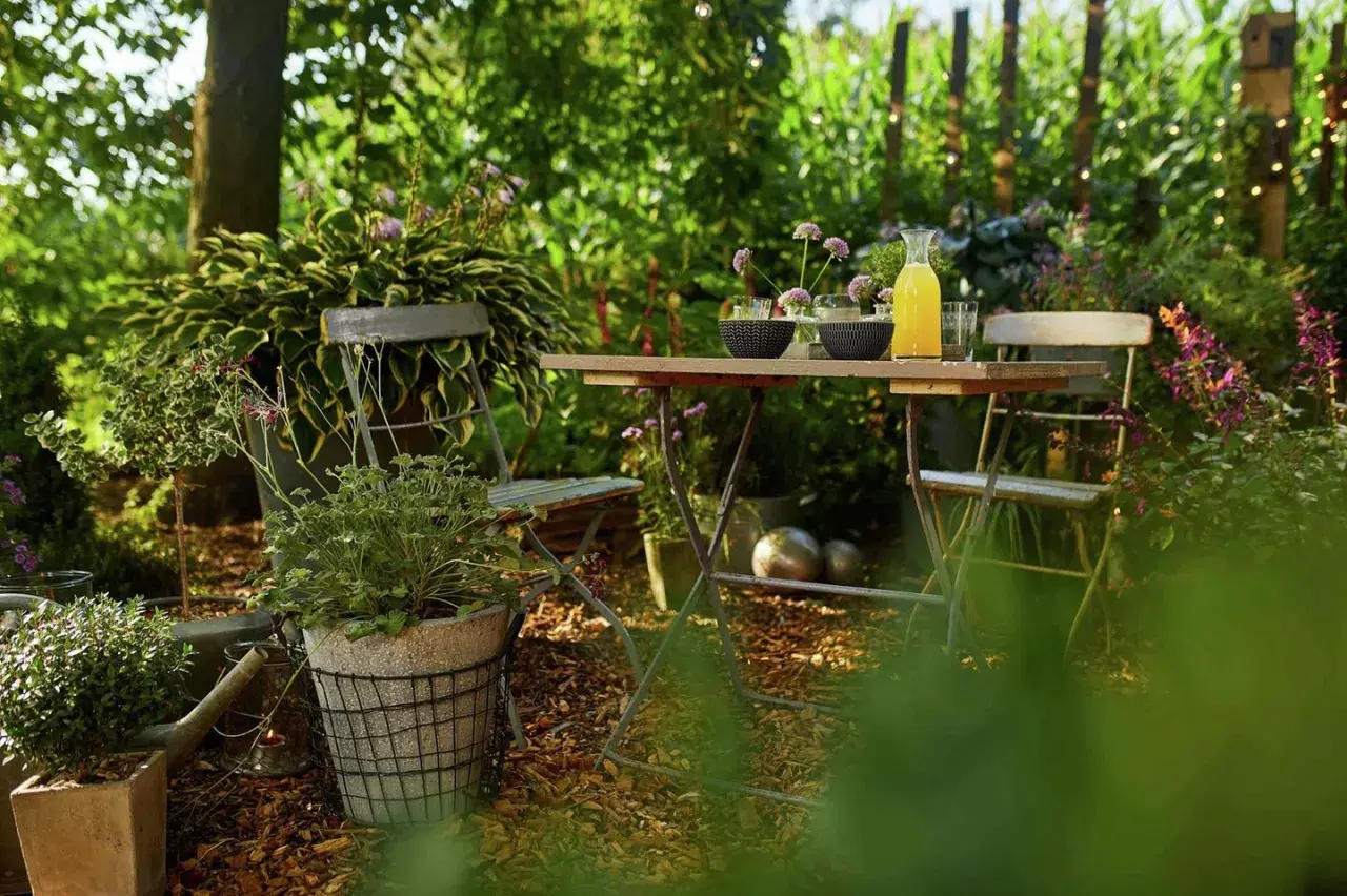 Gemütliche Gartenecke mit Tisch und Stühlen, umgeben von üppigem Grün. Rindenmulch bedeckt den Boden und bietet tolle Beet-Ideen für naturnahe Gestaltung.