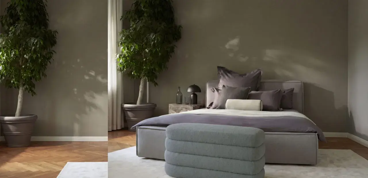 Ein stilvolles Schlafzimmer in gedeckten schlafzimmer farben mit zwei großen Pflanzen, einem Bett mit vielen Kissen und einem grauen Hocker.