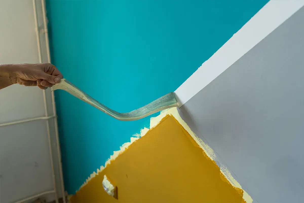 Hand zieht Klebeband ab, um eine gerade Kante zwischen türkisfarbener und grauer Wand zu schaffen. So gelingt das **richtig streichen**.