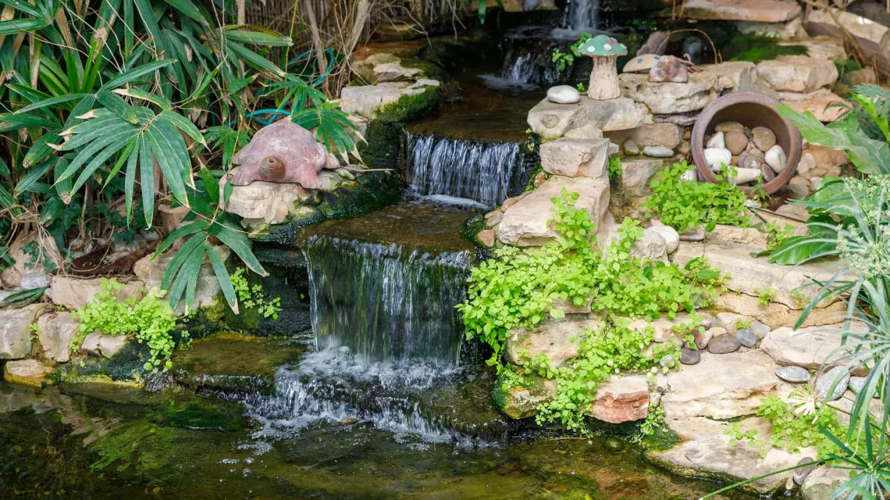 Ein kleiner Wasserfall plätschert in einen Teich, der mit Steinen und Pflanzen gestaltet ist. Eine Schildkrötenfigur wacht über das Wasser.