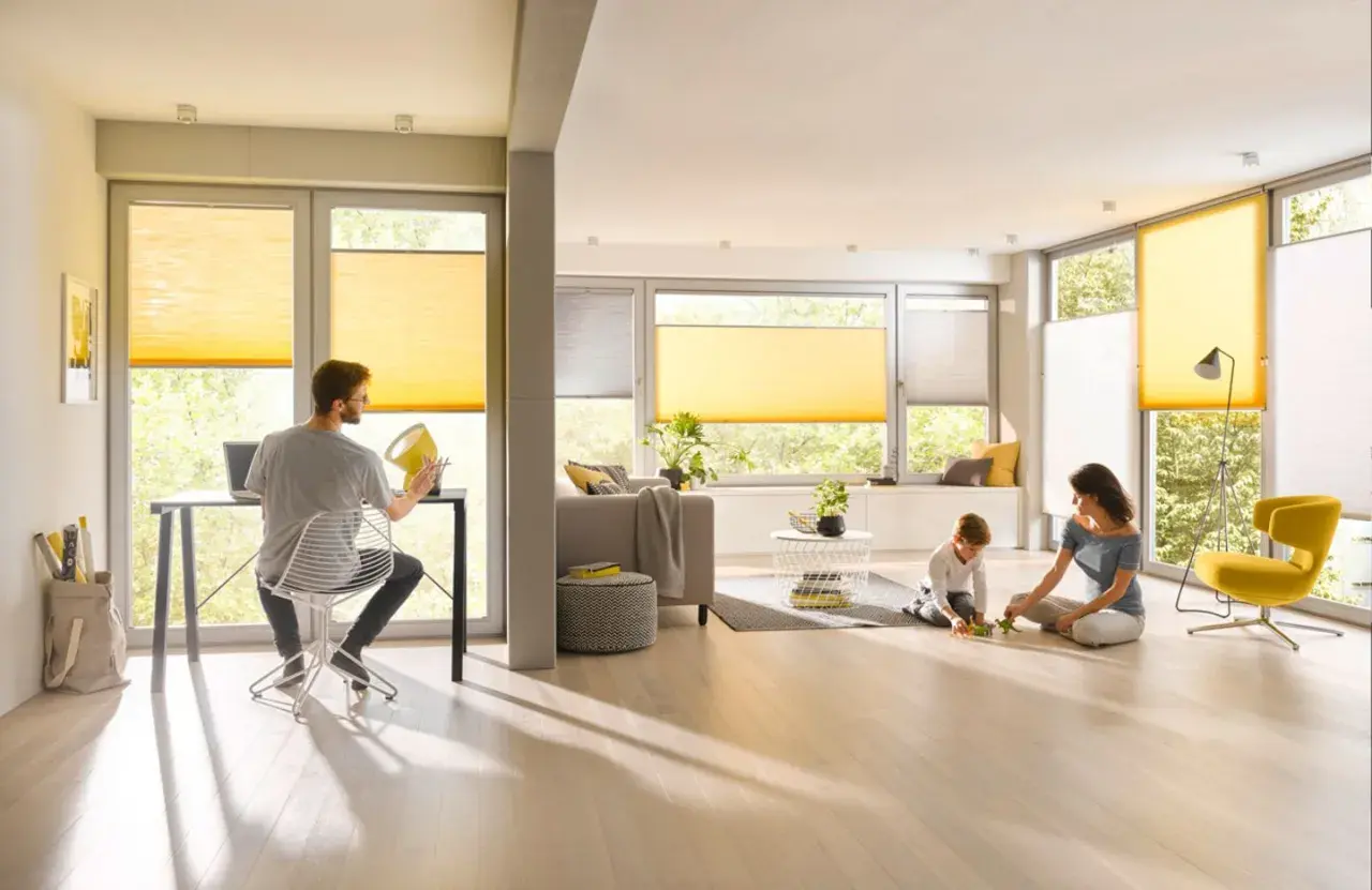 Familie genießt das Leben in einem hellen Wohnzimmer mit gelben Plissees, die das Fenster gestalten. Ein Mann arbeitet am Laptop, während Mutter und Kind spielen.