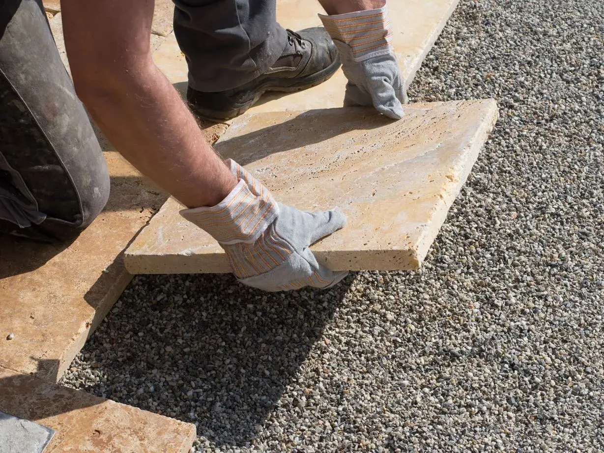 Arbeiter verlegt sorgfältig eine helle Terrassenplatte auf Kies.