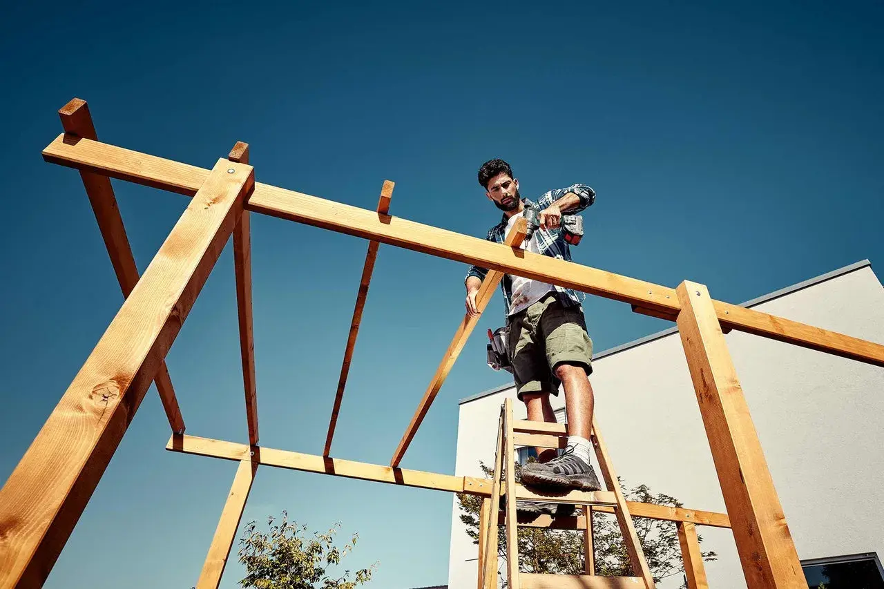 Mann auf Leiter schraubt Holzkonstruktion für ein Terrassendach selber bauen.