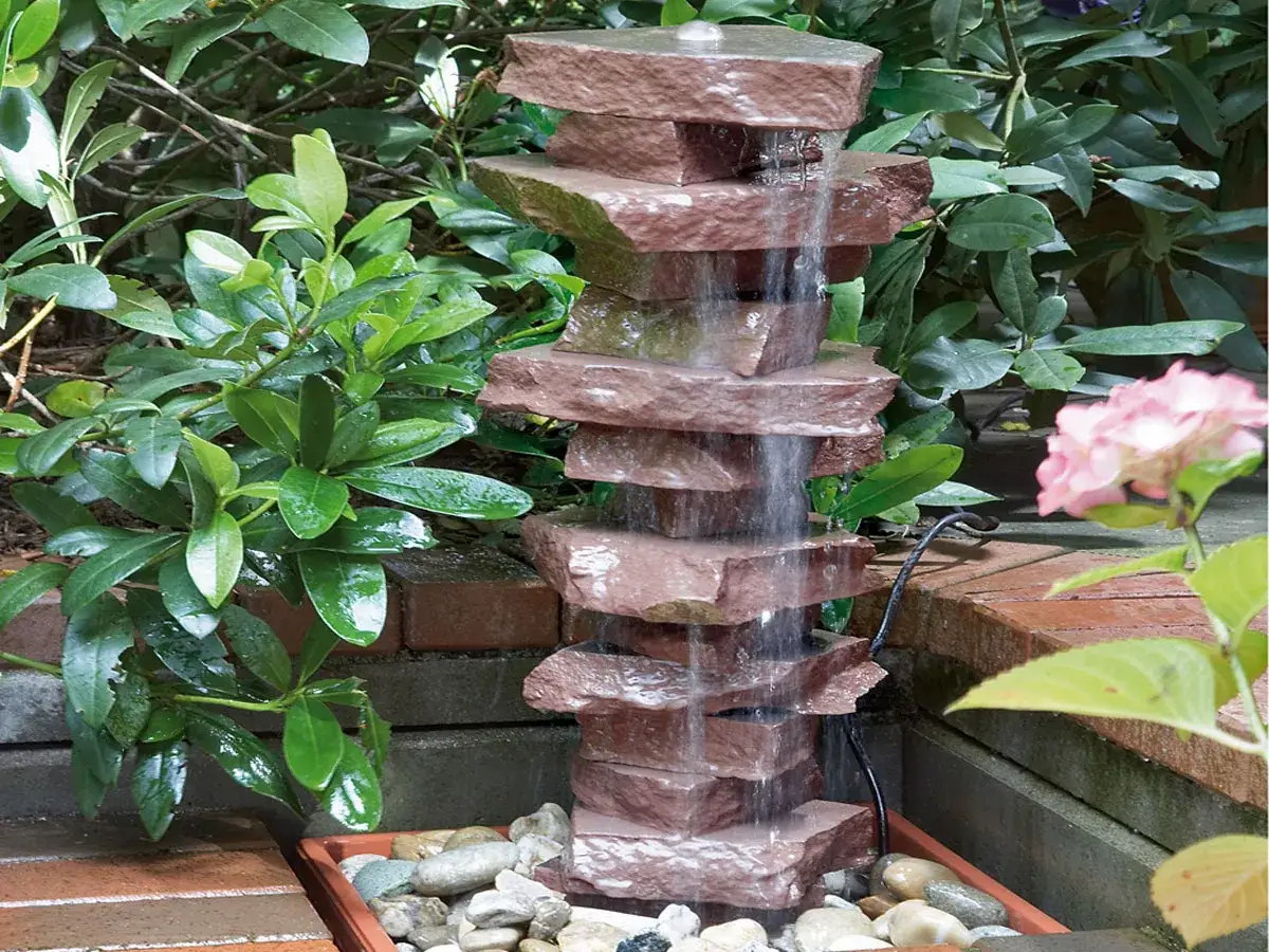 Ein moderner Brunnen Garten selber bauen mit gestapelten Steinen und pl&auml;tscherndem Wasser. Umgeben von gr&uuml;nen Pflanzen und einer rosa Bl&uuml;te.