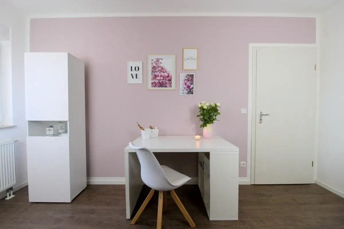 Ein wei&szlig;er Schreibtisch mit Stuhl und Blumenstrau&szlig; vor einer rosa Wand. Die Bilder an der Wand sind richtig gestrichen und schm&uuml;cken den Raum.