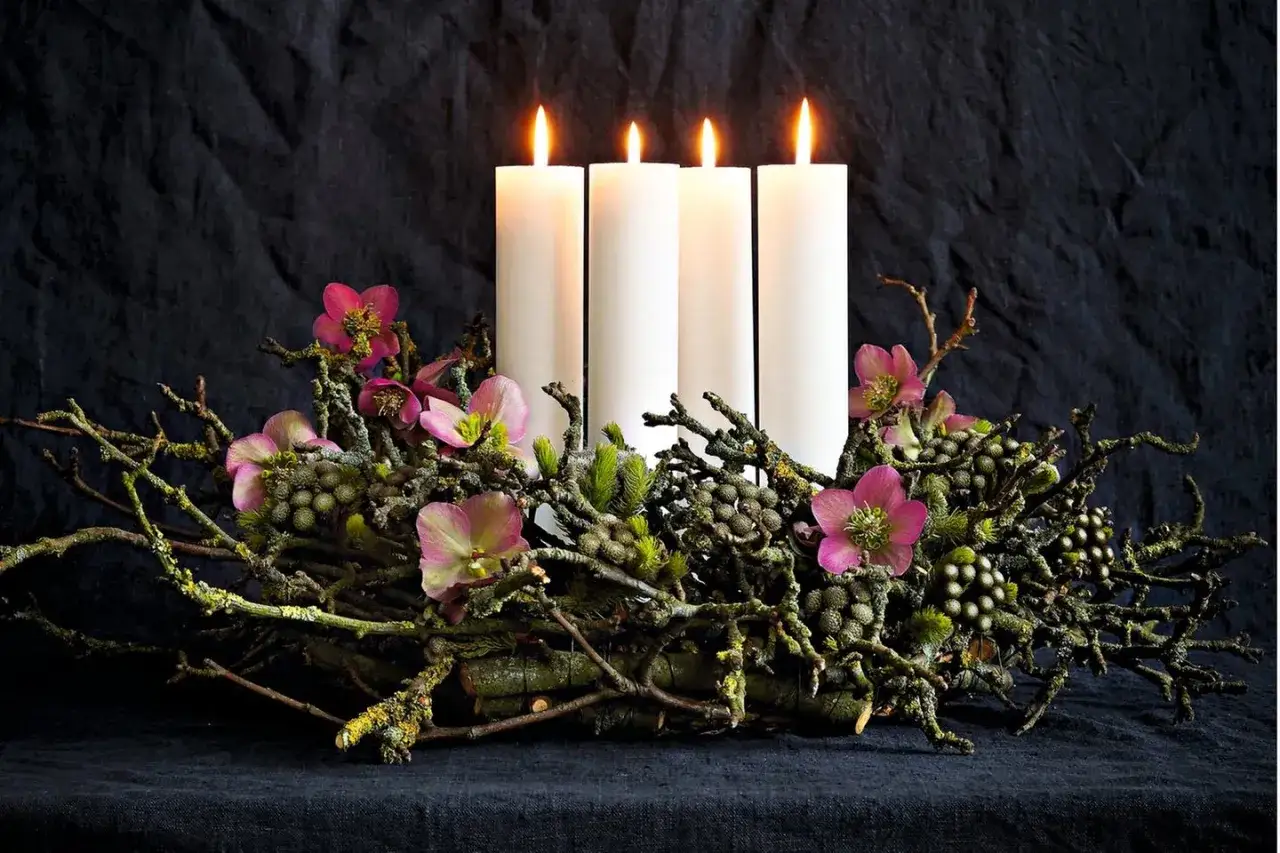 Ein adventskranz ausgefallen mit vier weißen Kerzen, umgeben von Ästen, rosa Christrosen und Zapfen auf dunklem Stoff.