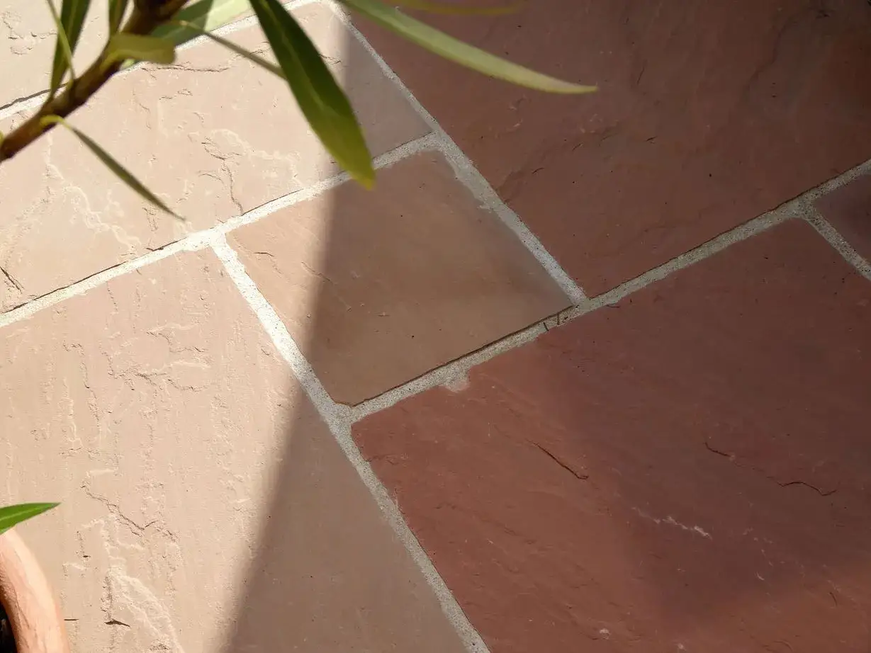 Terrasse verfugen: Nahaufnahme von Sandsteinplatten in Rot- und Beiget&ouml;nen, die mit hellem M&ouml;rtel verbunden sind. Ein Schatten f&auml;llt &uuml;ber die Fl&auml;che.