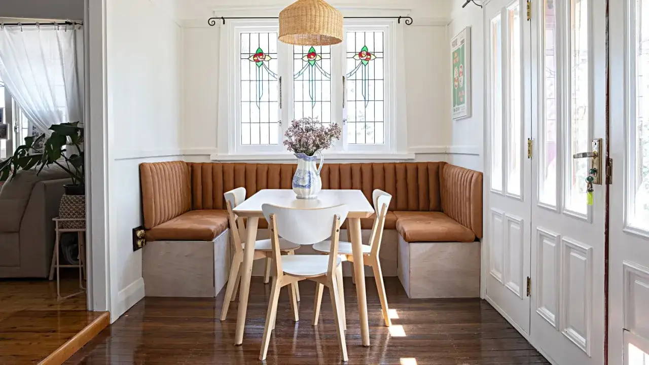Gem&uuml;tliches kleines Esszimmer einrichten mit brauner Eckbank, hellem Holztisch und wei&szlig;en St&uuml;hlen. Bunte Bleiglasfenster und Rattanlampe schaffen Atmosph&auml;re.