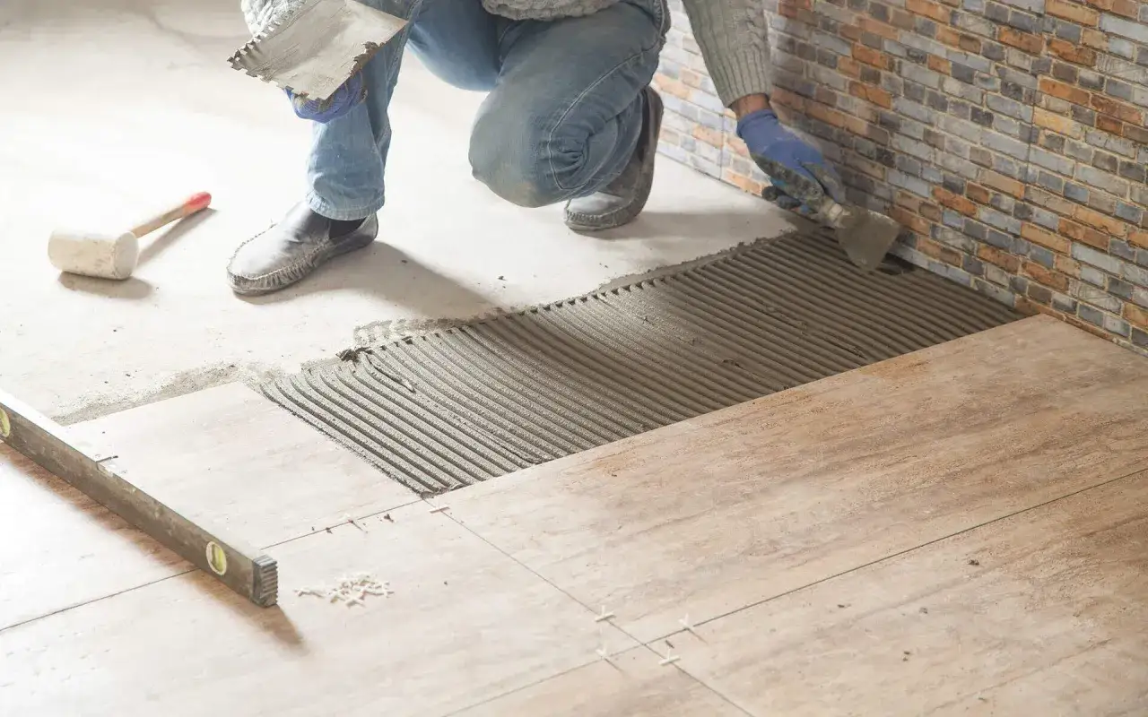 Hand klebt Fliesen auf Holz mit M&ouml;rtel. Werkzeuge wie Kelle und Wasserwaage liegen bereit.