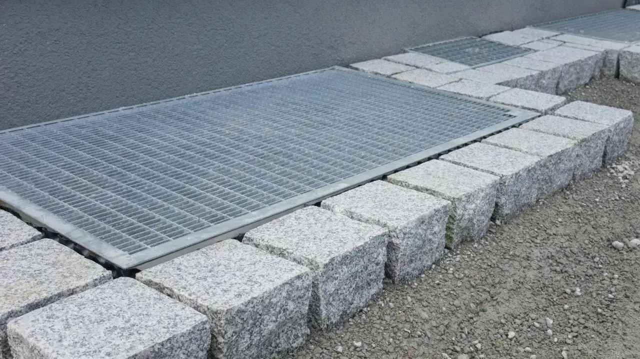 Metallgitter &uuml;ber einem Lichtschacht f&uuml;r ein Kellerfenster, flankiert von Granitw&uuml;rfeln und Schotter.