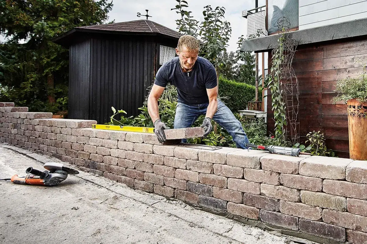 Mann baut eine Steinmauer im Garten. Werkzeuge wie Schleifmaschine und Wasserwaage liegen bereit.
