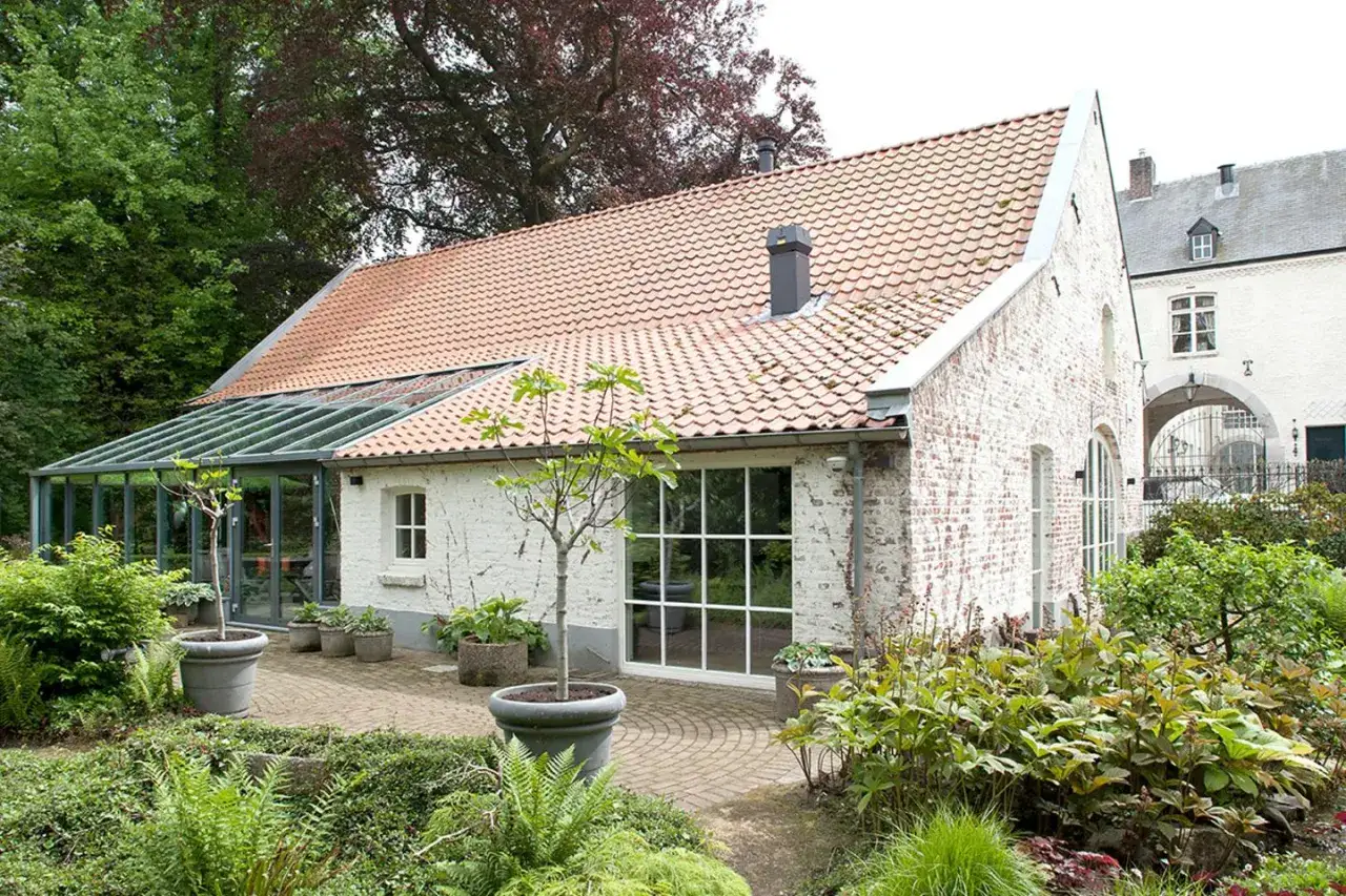 Moderne anbau wintergarten ideen: Glasanbau erweitert wei&szlig;es Backsteinhaus mit Ziegeldach, umgeben von &uuml;ppigem Gr&uuml;n und gepflasterter Terrasse.