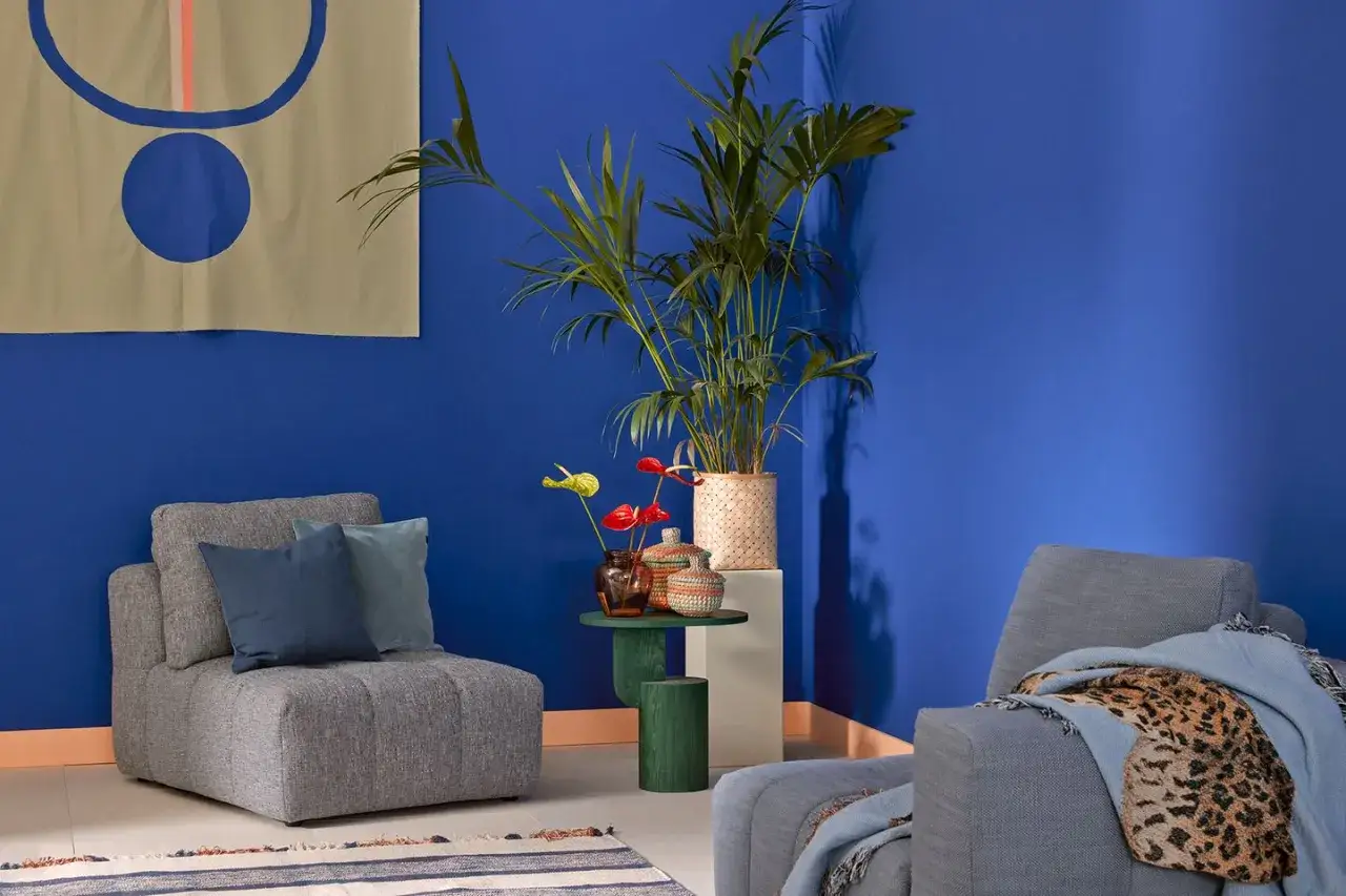 Ein gem&uuml;tliches Wohnzimmer mit blauen W&auml;nden. Graue Sessel, eine Palme und ein Teppich mit Streifen. Welche Farbe passt zu Blau? Hier: Orange und Beige.