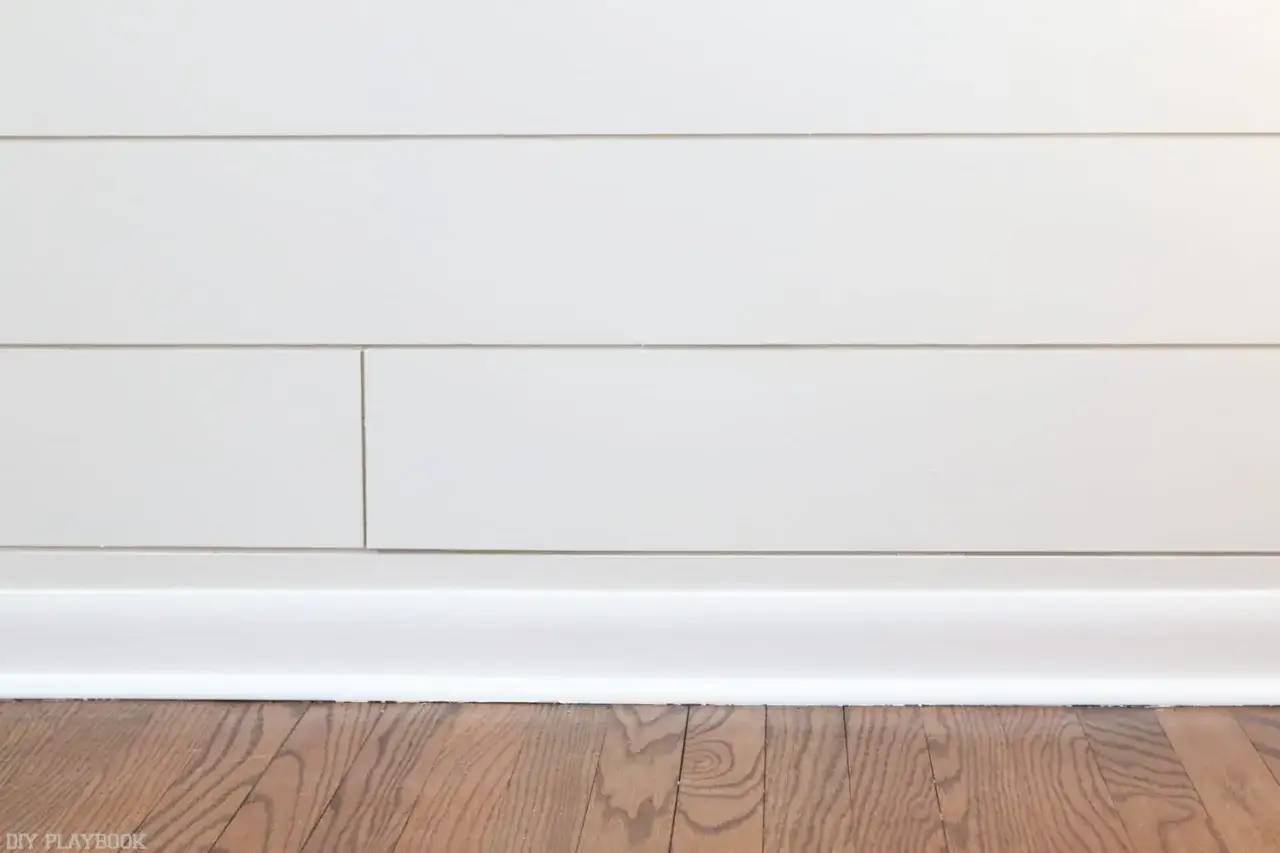 Weiße Sockelleisten werden an einer Wand mit horizontalen Paneelen befestigt. Der Holzboden ist im Vordergrund zu sehen.