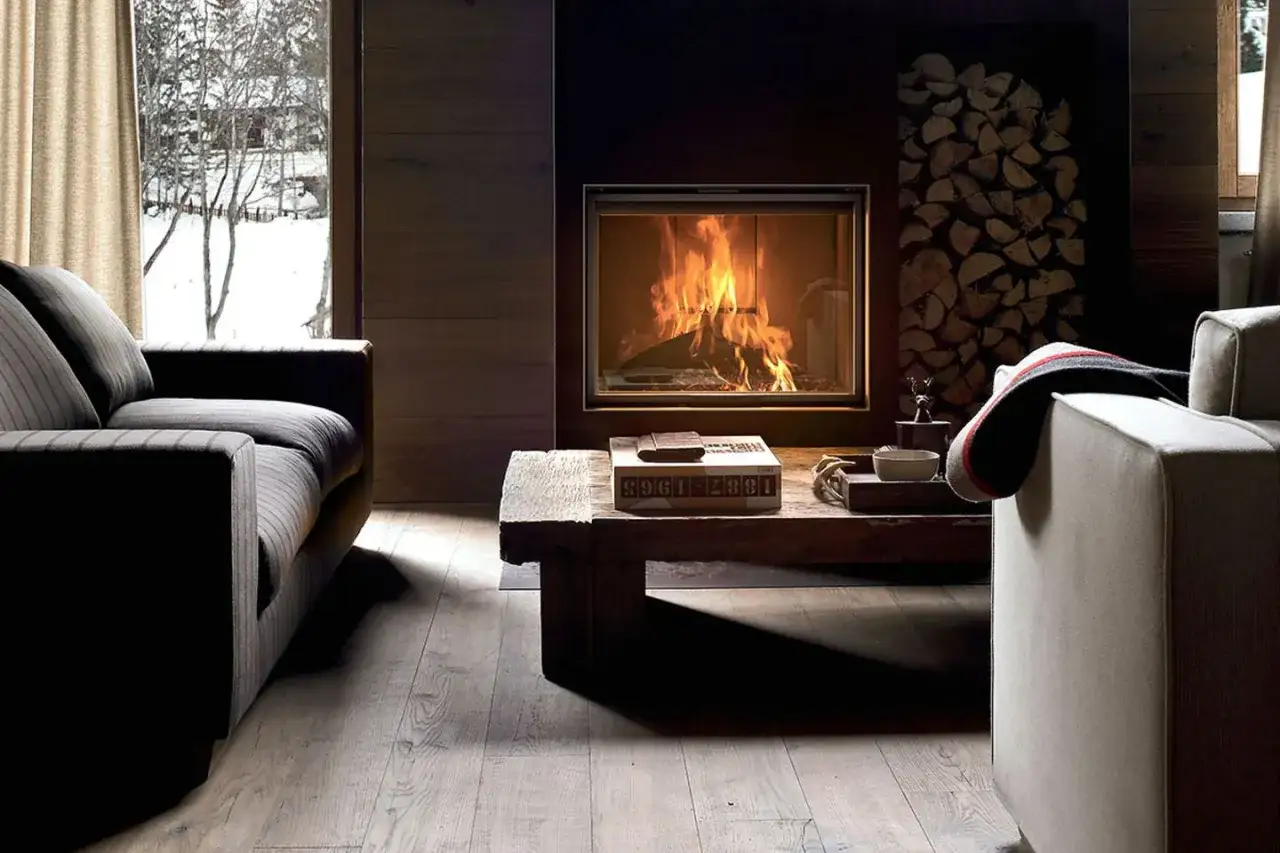 Gem&uuml;tliches kleines Kaminzimmer einrichten: Ein knisterndes Feuer im Kamin, zwei Sofas, ein Holztisch mit Deko und ein Fenster mit Winterblick.