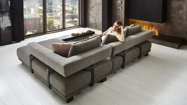 Moderne Wohnlandschaft mit grauem Stoffbezug, die als **Sofa als Raumteiler Ideen** dient. Eine Frau entspannt sich darauf, im Hintergrund ein Kaminfeuer.