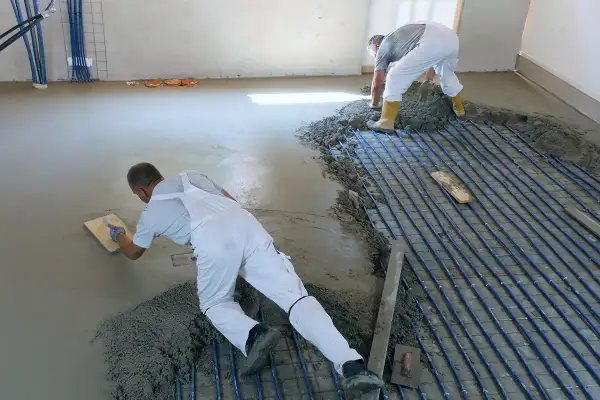 Zwei Männer machen Estrich selber, einer glättet den frischen Beton, der andere verteilt ihn über Heizungsrohren.