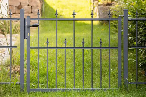 Graues Metallgartentor mit Griff und Schloss, bereit zum **Gartentor einbetonieren**. Dahinter grüner Rasen und eine Steinmauer.