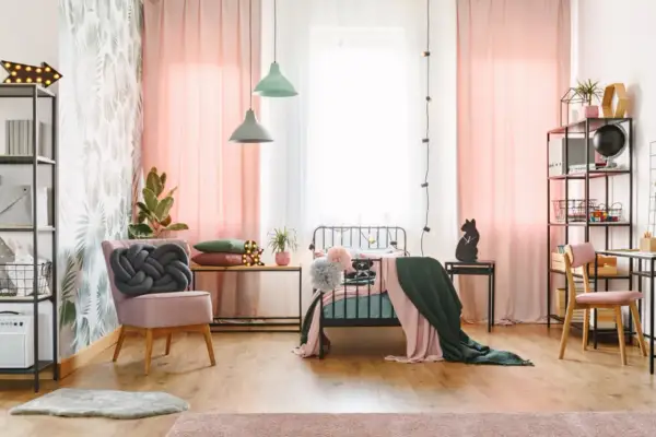 Ein stilvolles Jugendzimmer einrichten: Rosa Vorhänge, ein Bett mit Decken, ein Sessel mit Knoten-Kissen und ein Schreibtisch mit Regal.