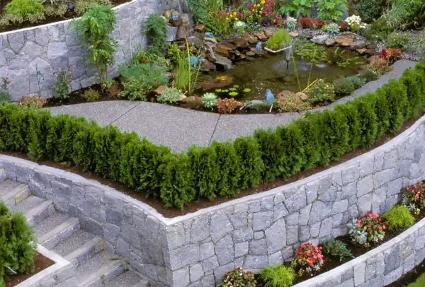 Eine malerische Gartentreppe führt an einer rustikalen steinmauer entlang, vorbei an einem kleinen Teich mit Wasserfall und üppiger Bepflanzung.