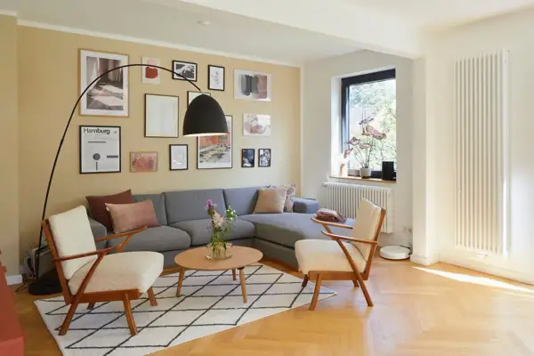 Gemütliches Wohnzimmer mit grauem Sofa, zwei Sesseln und einem Couchtisch. Die gelbe Wand ist mit vielen Bildern dekoriert, die den Rand des Raumes betonen.