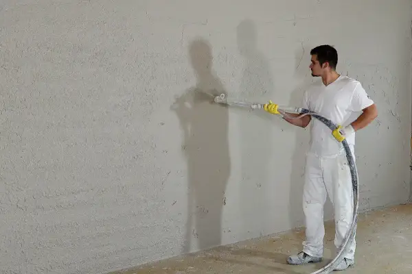 Mann verputzt eine Wand mit Spritzputz. So wird eine Wand verputzt: Gleichmäßige Bewegung der Spritzpistole.